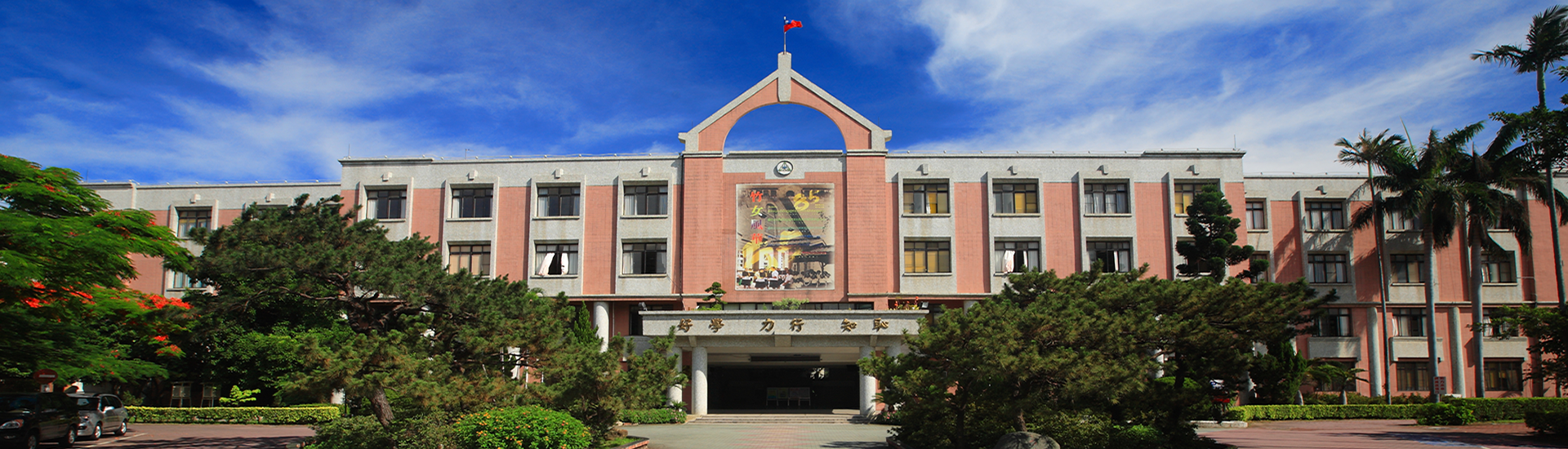 新竹女中校門口正面全景,前方有廣場、花圃與樹木,天空晴朗。